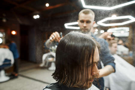 Young hipster guy with long hair getting new trendy haircut in the modern barbershop. Professional barber serving clientの写真素材