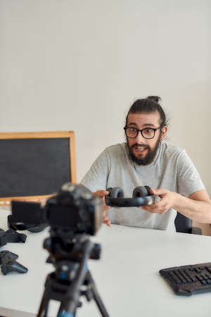Male technology blogger in glasses looking excited, holding headphones while recording video blog or vlog about new gadgets at home studioの写真素材