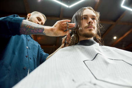 Young hipster guy sitting in barbershop chair and getting new trendy haircut while visiting modern...