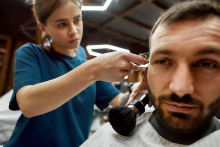 Close up shot of a professional barber girl or female hairdresser working with hair clipper, making trendy haircut for a handsome bearded manの写真素材
