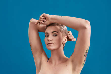 Beauty portrait of a young attractive half naked tattooed woman with perfect skin looking at camera, holding arms above her head isolated over blue backgroundの写真素材
