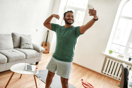 Be physically fit. Excited bearded active man holding smartphone, showing biceps while taking a...
