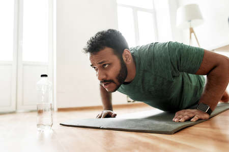 Young active man looking focused, exercising, doing push ups during morning workout at home. Sport,...