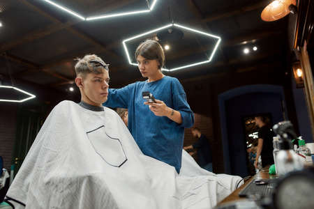 Side view of a young handsome guy sitting in barber shop chair while professional barber girl working with hair clipper, making trendy haircut for himの写真素材