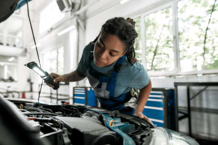 Moments of caring. Young african american woman, professional female mechanic looking, examining under hood of car with torch at auto repair shopの写真素材