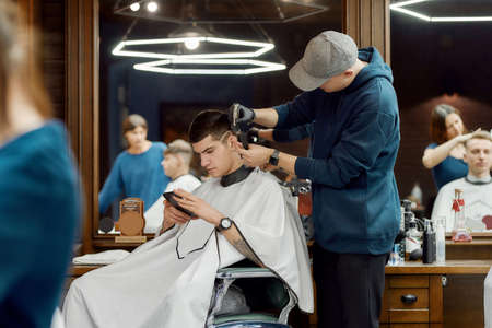 Side view of a professional barber working with hair clipper, making trendy haircut for client sitting in barber shop chair and using his smartphoneの写真素材