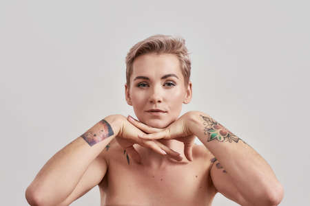 Portrait of half naked tattooed woman with short hair looking at camera, posing with hands under her...