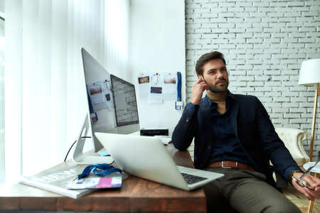 Discussing business. Young confident businessman or sales manager wearing wireless earphones talking with client or partner while working on computer in the modern officeの写真素材