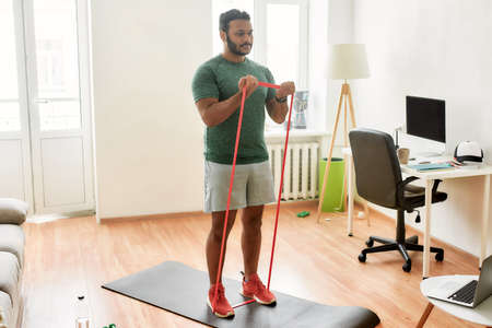 Tone up your body. Full length shot of young active man watching online video training on laptop, exercising with resistance band during morning workout at homeの写真素材