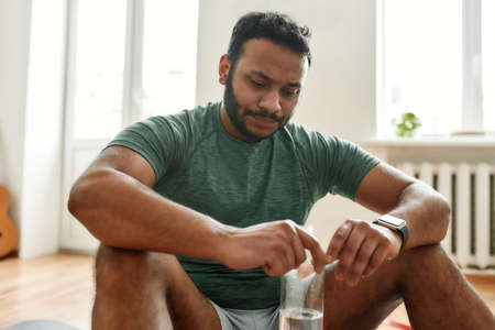Exhausted. Young tired man looking down dissatisfied, holding water bottle in his hands while resting after training workout at home. Sport, healthy lifestyleの写真素材
