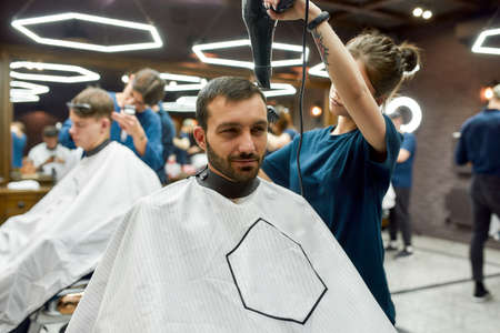 Handsome man visiting barber shop. Professional young barber girl drying hair of a handsome bearded man, serving clientの写真素材