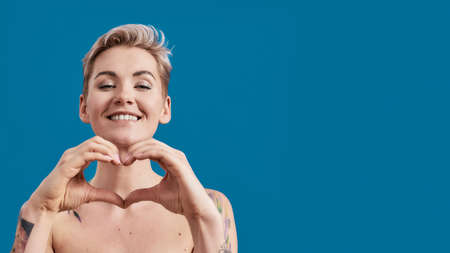 Portrait of a young attractive half naked tattooed woman with perfect skin looking happy, showing heart with hands isolated over blue backgroundの写真素材