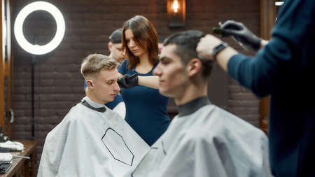 People in barbershop. Side view of a young focused barber girl or female hairdresser making new trendy haircut for clientの写真素材