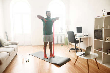 Healthy life. Full length shot of male fitness instructor showing exercises with resistance band while streaming, broadcasting video lesson on training at home using laptopの写真素材