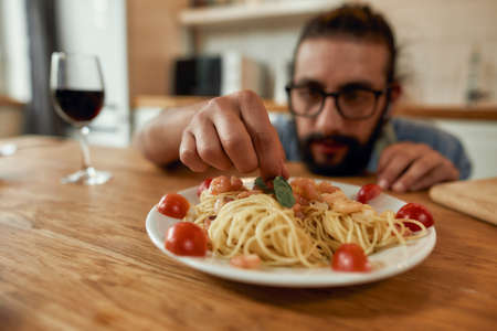 Great food. Young man, cook serving garlic butter shrimp pasta, decorating plate with basil and tomatoes. Cooking at home, Mediterranean cuisine conceptの写真素材
