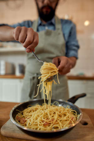 Cropped shot of man in apron, cook holding spaghetti on fork while preparing garlic butter shrimp pasta at home. Mediterranean cuisine conceptの写真素材