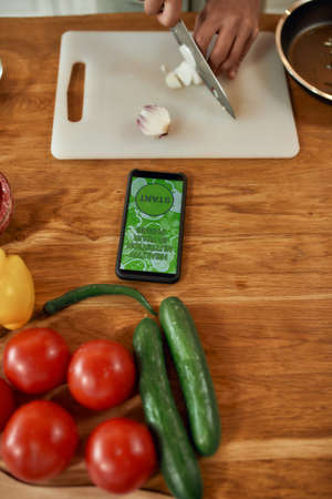 Close up of phone and vegetables on the table. Man cutting onion on chopping board, using smartphone app while cooking dinner. Healthy nutrition, technology conceptの写真素材