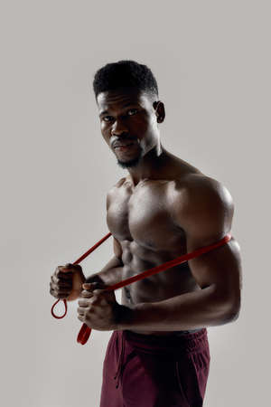 Young muscular african american man looking at camera, while holding resistance band isolated over...