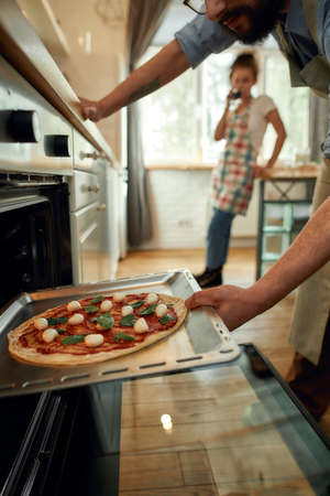 Professional cook making pizza at home. Man putting raw pizza in modern oven for baking. Woman standing in the background. Hobby, lifestyleの写真素材