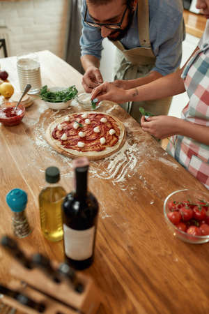 For a tasty pizza. Young couple making pizza together at home. Man in apron, professional cook adding mozzarella cheese on the dough while woman helping him adding basil. Hobby, lifestyleの写真素材