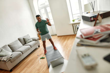Hi guys. Full length shot of male fitness instructor greeting, waving hand while streaming,...