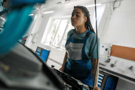 Young african american woman, professional female mechanic opening car hood and checking car at auto repair shopの写真素材