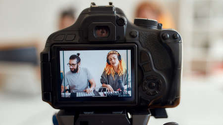 Woman with dreadlocks singing and playing. Female and male blogger recording making music using synthesizer, drum pad machine. Focus on camera screen