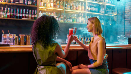 Have a nice night. Two women having fun, toasting, drinking cocktails while sitting at the bar...