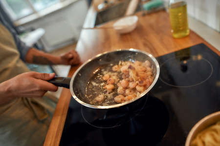 Close up of hand of man tossing shrimp with garlic and onion in the frying pan. Cook preparing dish with seafood. Mediterranean cuisine conceptの写真素材