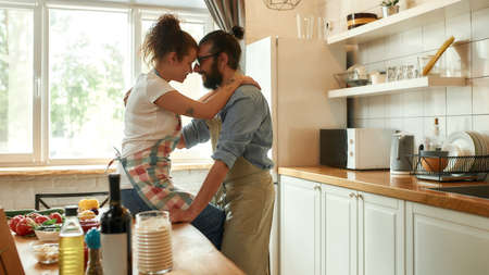 Young man looking at his girlfriend, hugging while standing in the kitchen. Couple embracing while making pizza with vegetables indoors. Cooking together, hobby, lifestyleの写真素材