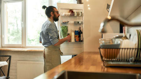 Young man, professional cook in apron taking ingredients out of the fridge while getting ready to prepare a meal, standing in the kitchen. Cooking at home conceptの写真素材