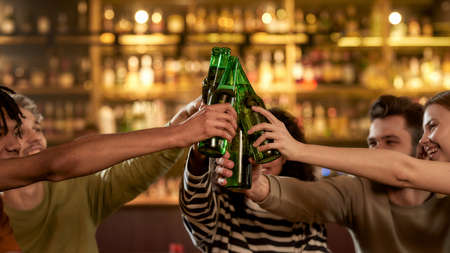 Close up of hands of people in the bar clinking bottles while drinking beer together. People,...