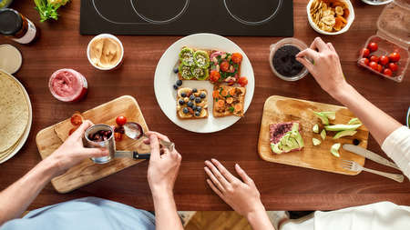 Top view of couple of vegetarians preparing various healthy toasts in the kitchen together. Man getting out sun dried tomatoes while woman adding chia seedsの写真素材