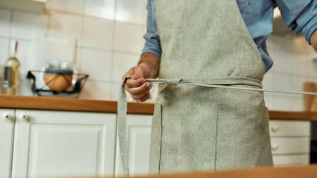 Close up of young man, professional cook tying his apron while getting ready to prepare a meal,...