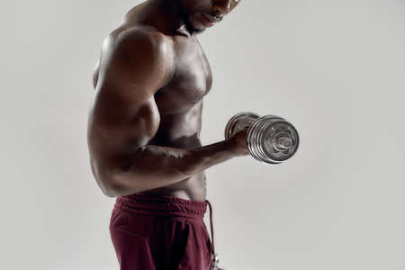 Cropped shot of young muscular african american man lifting weights, standing isolated over grey background. Sports, workout, bodybuilding conceptの写真素材