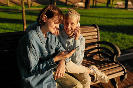 Two joyful girls using smartphone, sitting on the bench in the city park. Lesbian couple holding...