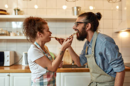 Enjoy the taste. Young man and woman in apron feeding freshly baked pizza to each other while standing in the kitchen. Love, relationships conceptの写真素材