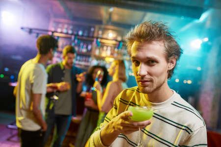 Portrait of caucasian young man looking at camera while posing with a cocktail in his hand and...