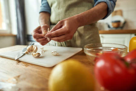 Close up of hands of man in apron peeling garlic while preparing healthy meal, soup in the kitchen. Cooking at home, Italian cuisine conceptの写真素材