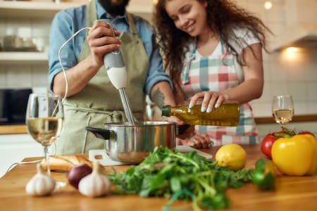 Cropped shot of man, chef cook using hand blender while preparing a meal. Young woman, girlfriend in apron pouring olive oil in the pot, helping him in the kitchenの写真素材