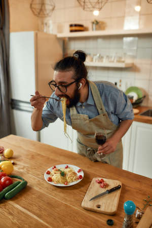 Tasting meal. Young man in apron, cook holding spaghetti wrapped around a fork and a glass of wine while eating pasta in the kitchen at home. Italian cooking conceptの写真素材