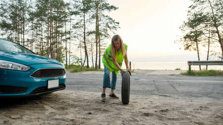 Full length shot of attractive young woman wearing reflective vest, rolling spare tire to change the...