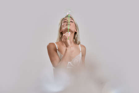 Sensual young woman in sexy lingerie having fun while blowing soap bubbles at camera, posing isolated over grey backgroundの写真素材