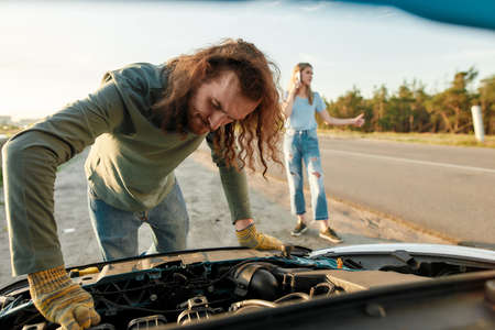 Man looking under the hood of broken car, girl calling car service or assistance in the backgroundの写真素材