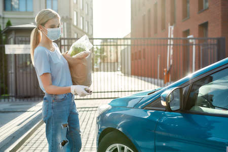 Young woman in medical mask and protective gloves presses unlock button on car remote control, unlocking door alarm system while holding grocery bagの写真素材