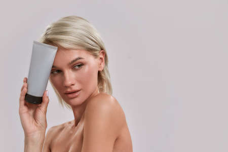 Portrait of young woman with perfect fresh skin looking aside, holding and advertising cosmetic...