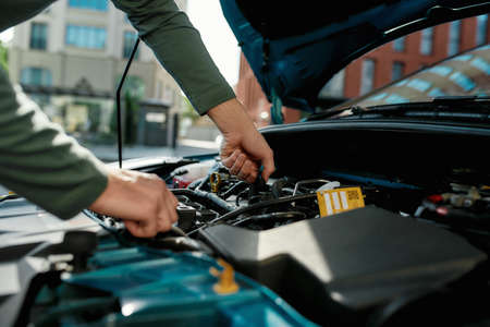 Close up of hands of young man examining broken down car engine, standing near his car with open...
