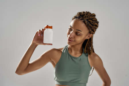 Healthy products for sportsmen. Young mixed race fitness woman in sportswear holding bottle with sports nutrition supplements while standing isolated over grey backgroundの写真素材