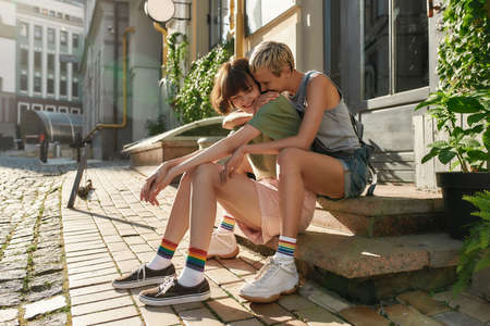Full length shot of young lesbian couple, two girls hugging and smiling, sitting on the steps while having a walk around the city. LGBT, relationship conceptの写真素材