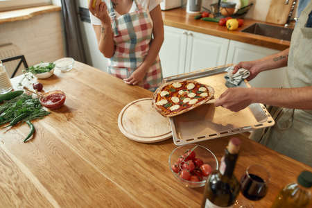 Fresh out of the oven. Young man in apron, cook taking tray with hot freshly baked pizza out of oven. Couple making pizza together. Hobby, lifestyleの写真素材
